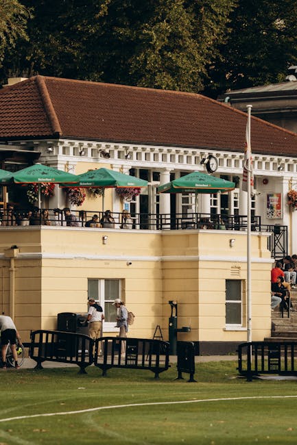 A two-storey garden maintenance facility situated in a public outdoor area, featuring a cream-colored facade with decorative architectural elements and a red-tiled roof. The upper terrace is shaded by